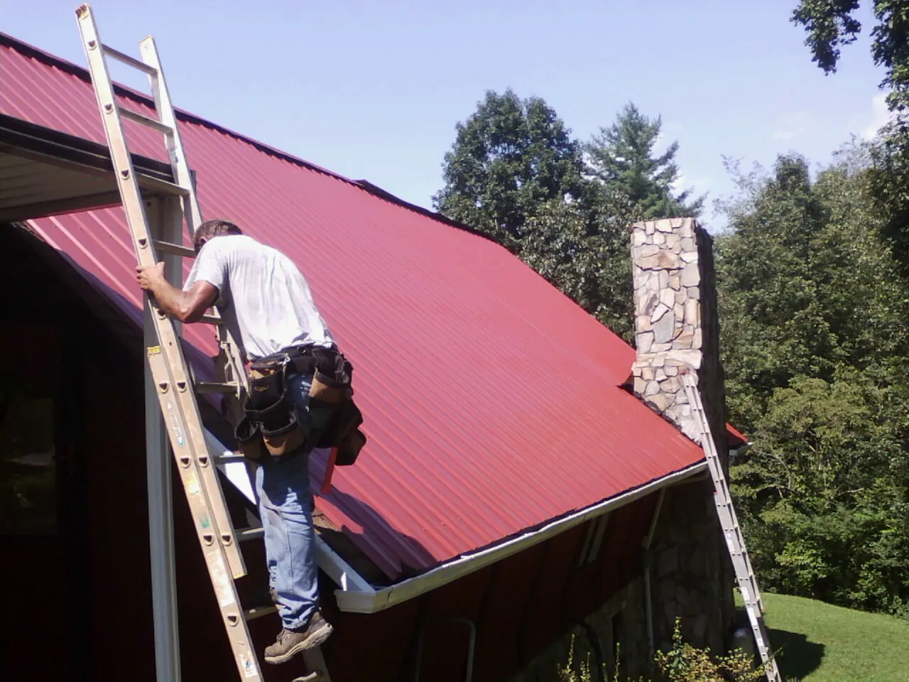 Roofer on a ladder beside a red metal roof and stone chimney — Soffit & Fascia Repair by Roofing Brothers in Jonesborough